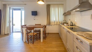 Une cuisine avec des meubles de rangement en bois, une table à manger avec des chaises, un téléviseur fixé au mur et une fenêtre avec des rideaux.