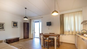 Une pièce comprenant une table à manger en bois, des chaises, un canapé, un téléviseur fixé au mur et un coin cuisine avec un évier et un plan de travail.