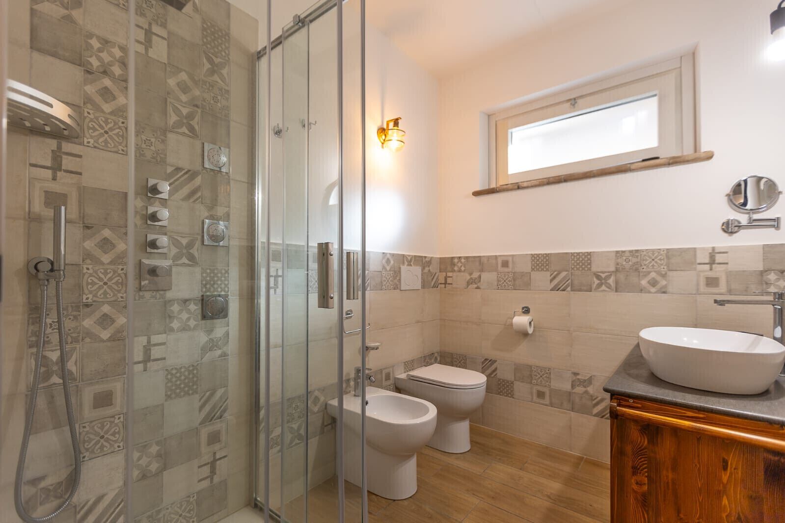 A bathroom with a glass-enclosed shower, two toilets, a sink, and patterned tiles.