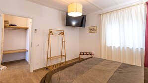 A bedroom with a bed, a wooden wardrobe, a hanging rack, a television, and a window with curtains. - Le Vigne - Appartamento Palazzo Chiavoni (Bolsena)