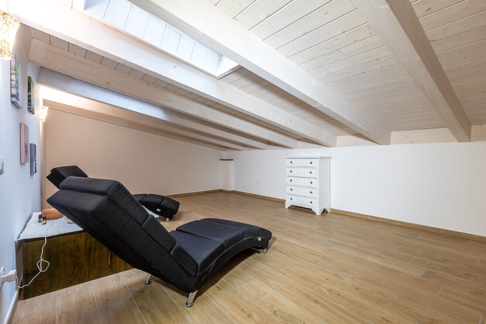 Een ruime kamer met een schuine kap, een massagetafel, een klein kastje en een raam.