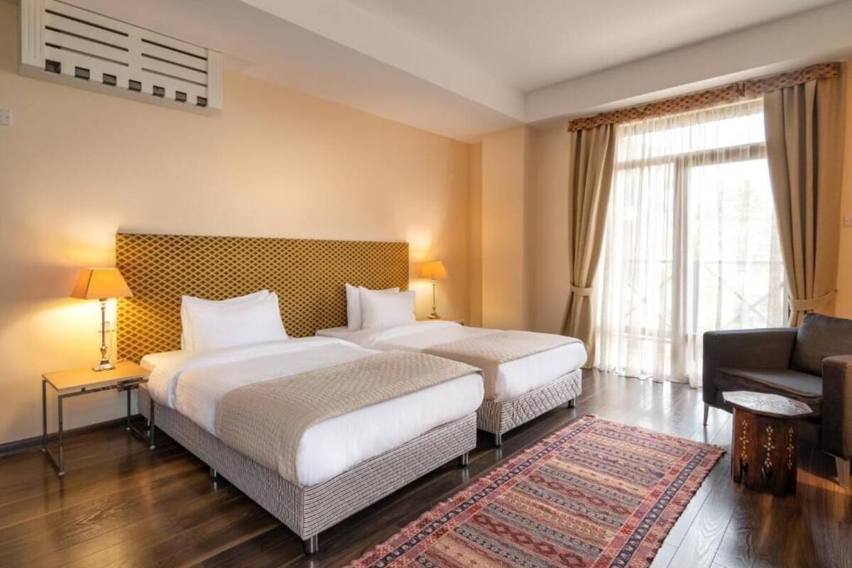 A hotel room with two beds, a patterned headboard, a large window with curtains, a patterned rug, and a small round table.