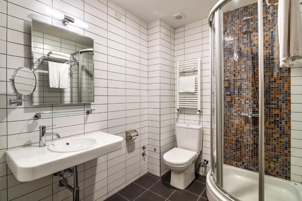 Casa de banho com lavatório branco, sanita, chuveiro com porta de vidro e parede revestida a azulejos.