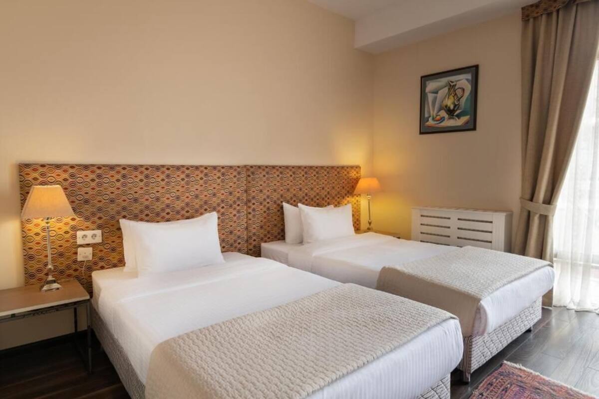 A hotel room with two beds, a patterned headboard, bedside lamps, a painting on the wall, and a window with curtains.