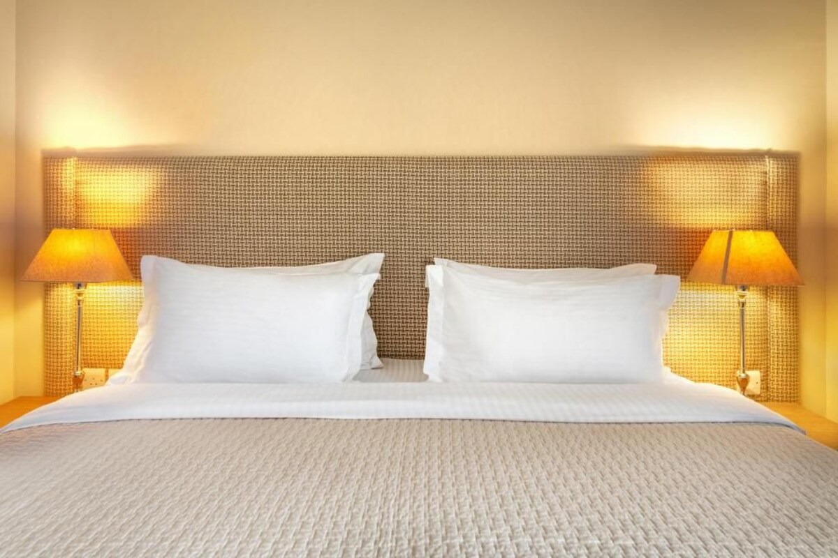 Quarto de hotel com uma cama grande, duas luminárias de cabeceira e uma cabeceira texturizada.