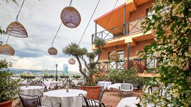 An outdoor patio with round tables and chairs, a brick building, and hanging wicker lamps. - Ginger Hotel (Tbilisi)