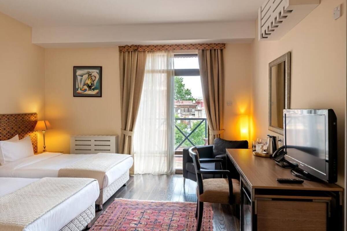 A hotel room with two beds, a desk with a computer, a TV, and a balcony with curtains.