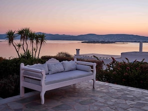 A white outdoor sofa with striped cushions, a stone patio, and a view of a body of water and distant hills. - Seafront Villa Elia -island Living- (Paros)