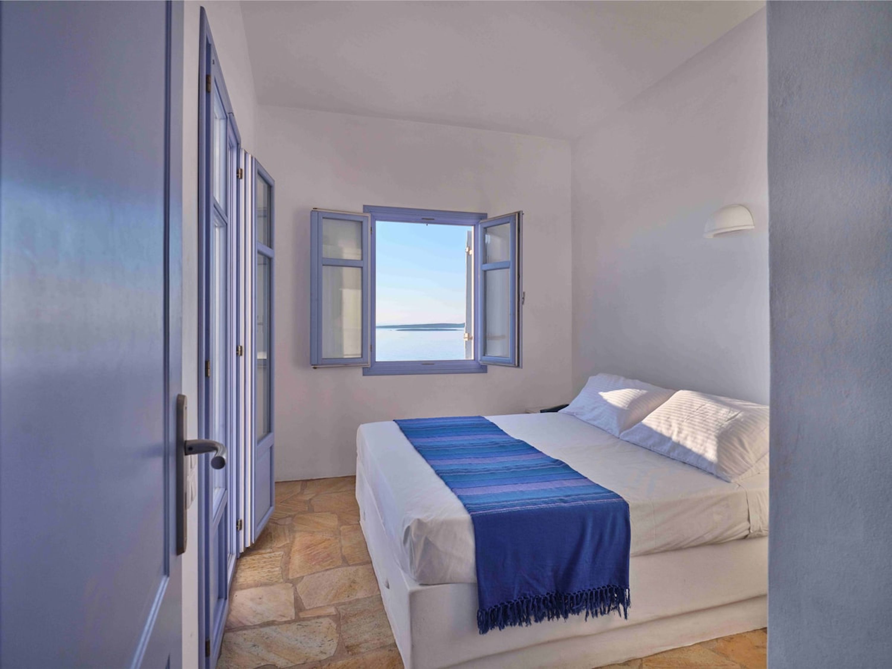 Ein Schlafzimmer mit Bett, Fenster mit geöffnetem Klappladen und Steinboden.