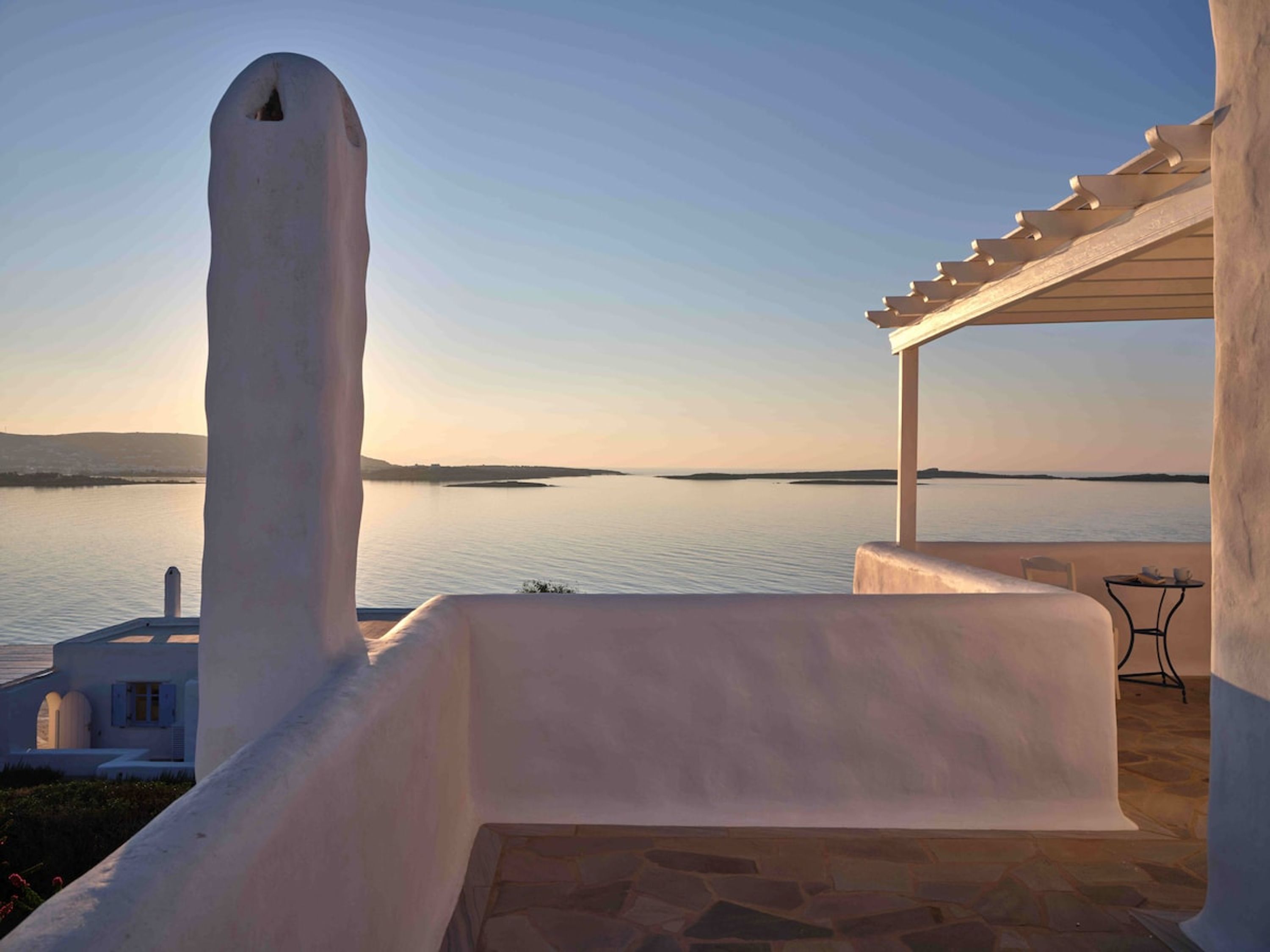 A white building with a stone floor, a pergola, and a view of a calm body of water.