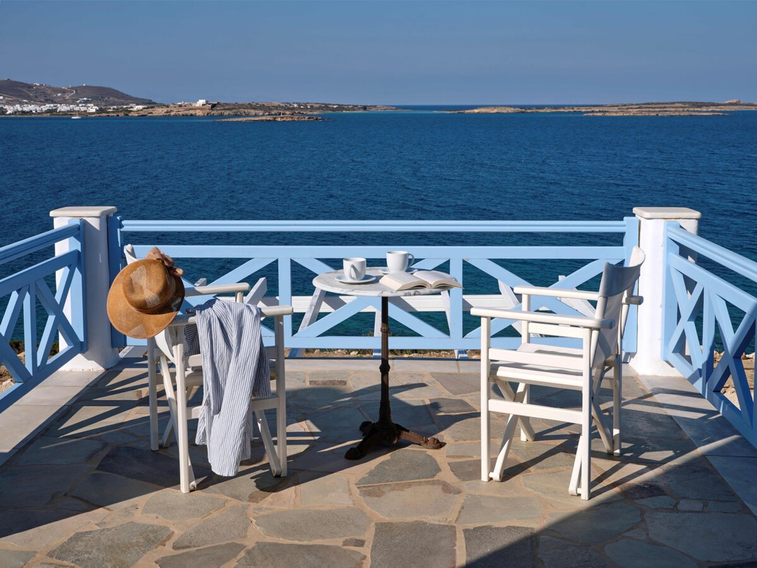 Eine Seeterrasse mit weißen Stühlen, einem Tisch und einem Hut, mit Blick auf das Meer.
