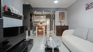 A modern living room with a sofa, a TV, a dining area with chairs, and a kitchen in the background. - Sweet Home by RentalsPro - Nea Moudania (Nea Moudania)