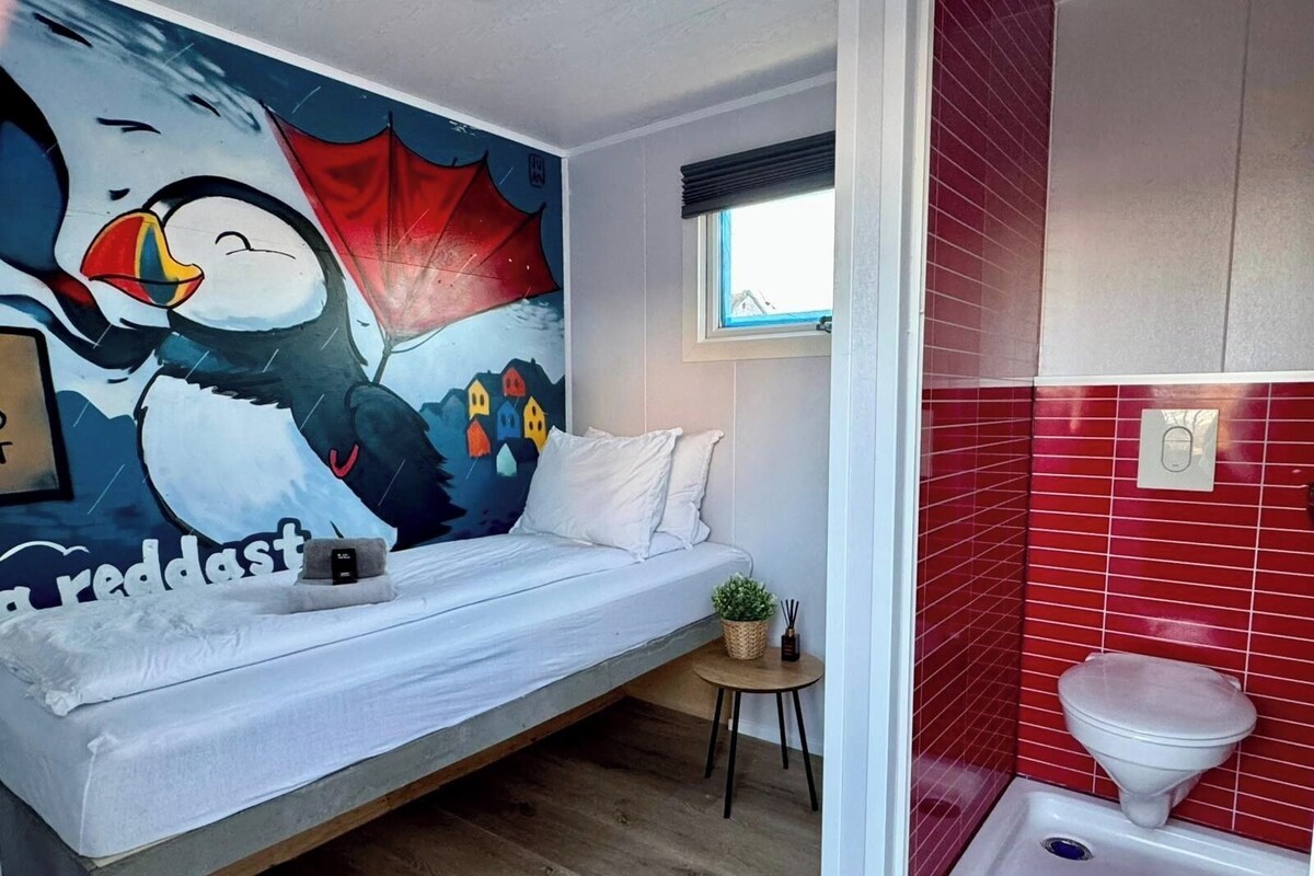 Un dormitorio con cama, una mesita con una planta, un mural en la pared y un baño con piso de baldosas rojas.