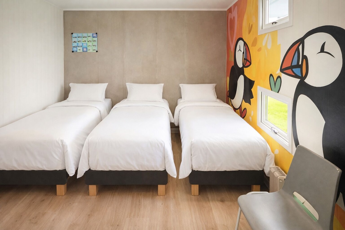Habitación con tres camas individuales, suelo de madera, pared con un mural de pingüinos coloridos y una ventana pequeña.