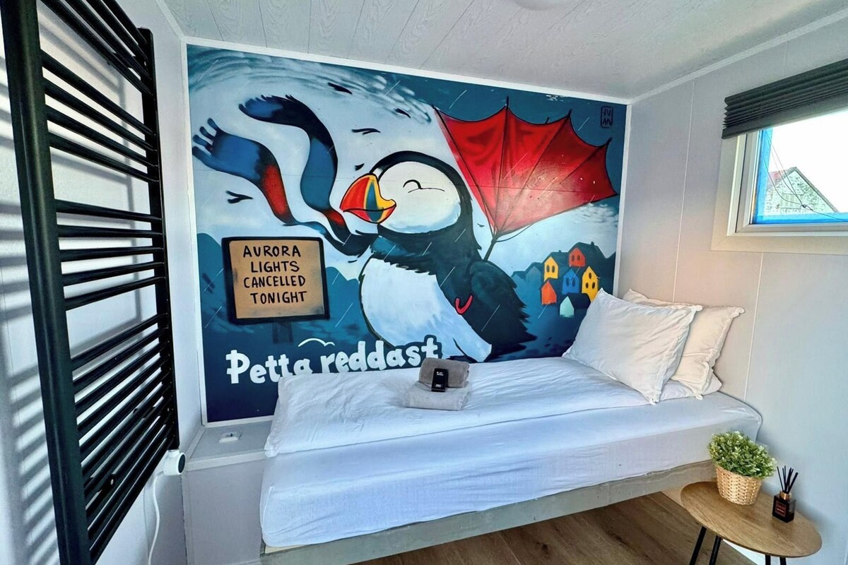 A bedroom with a mural of a penguin, a bed, a bedside table, and a window.