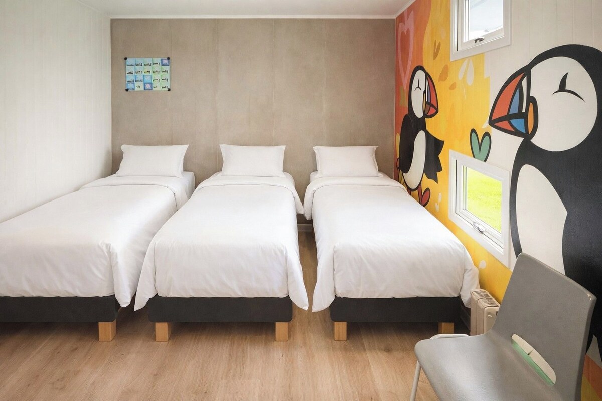 A room with three single beds, a wooden floor, a wall with a colorful mural, and a window.