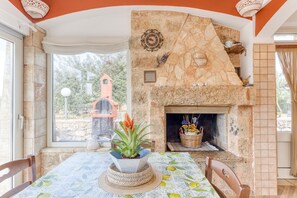 Un comedor con chimenea de piedra, una mesa con mantel floral y vista a una construcción de techo rojo en el exterior.