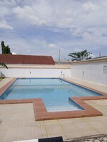 Piscina retangular com deck de madeira e uma parede branca com telhado marrom ao fundo.