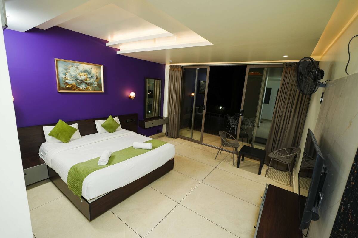 A modern hotel room with a large bed, a painting on the wall, a sliding glass door to a balcony, and a TV mounted on the wall.