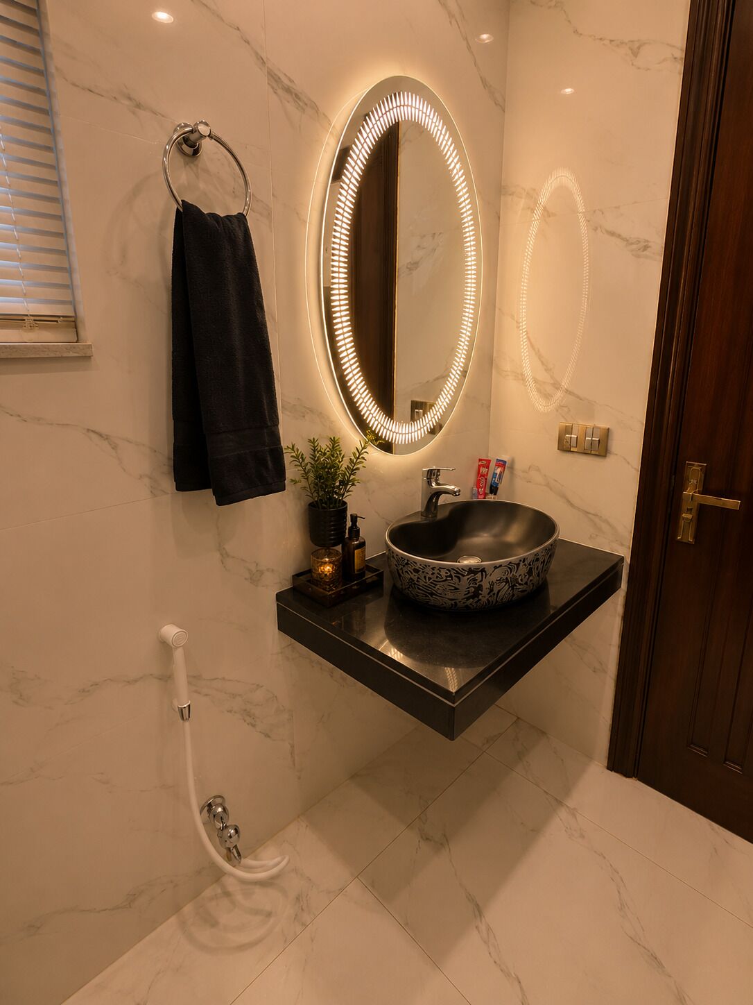 Une salle de bain avec un mur en marbre, une serviette noire, un miroir rond avec éclairage, un lavabo et une douche.