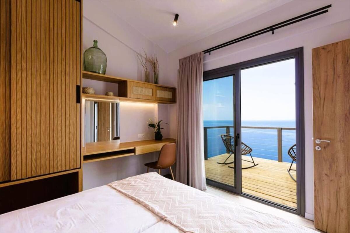 A bedroom with a bed, a desk, a chair, a large window, and a balcony with chairs.