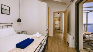 A hotel room with a bed, a view of the sea, a hallway leading to a balcony, and a wooden floor. - Anemos Luxury Villas (Agios Vasileios)