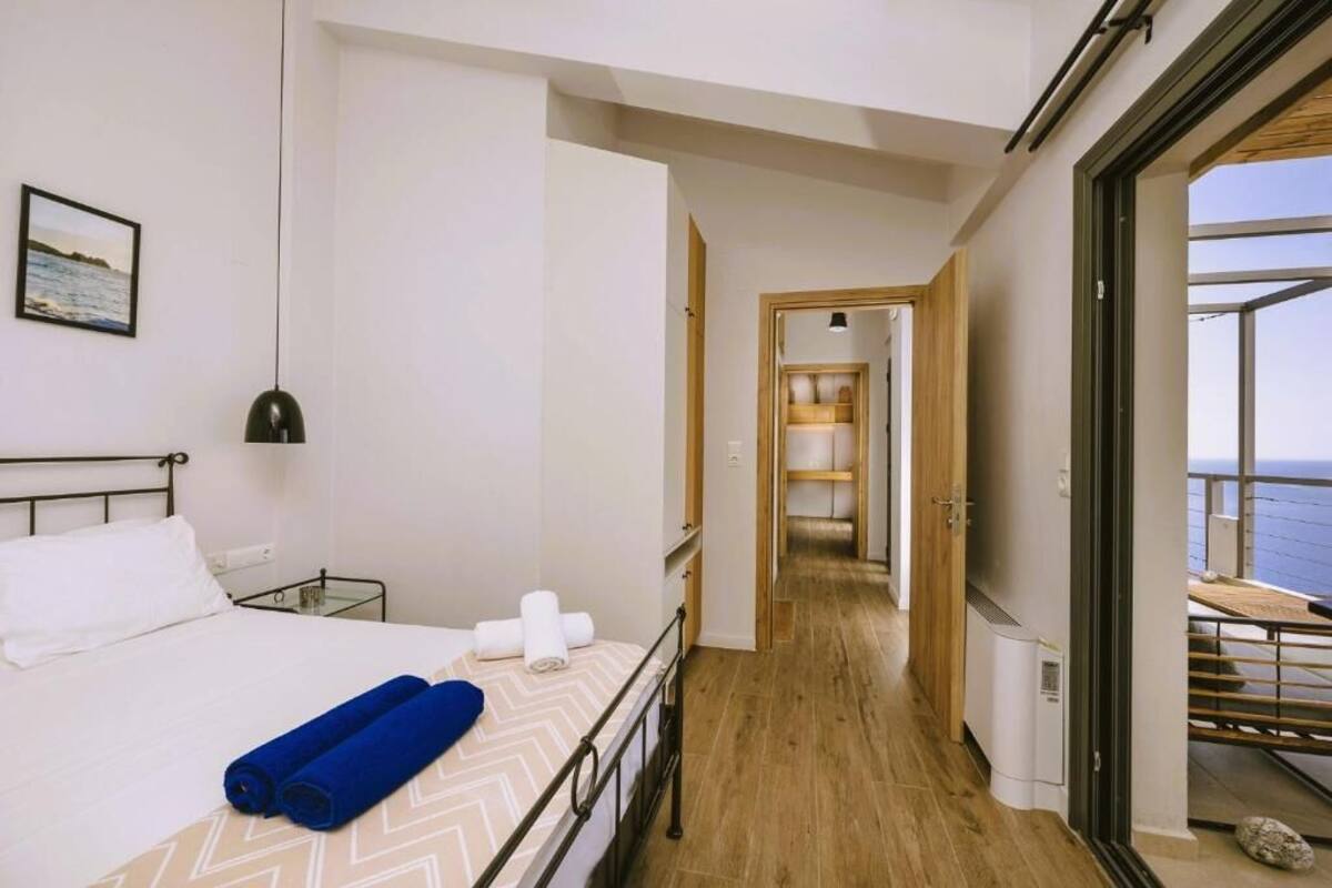 A hotel room with a bed, a view of the sea, a hallway leading to a balcony, and a wooden floor.