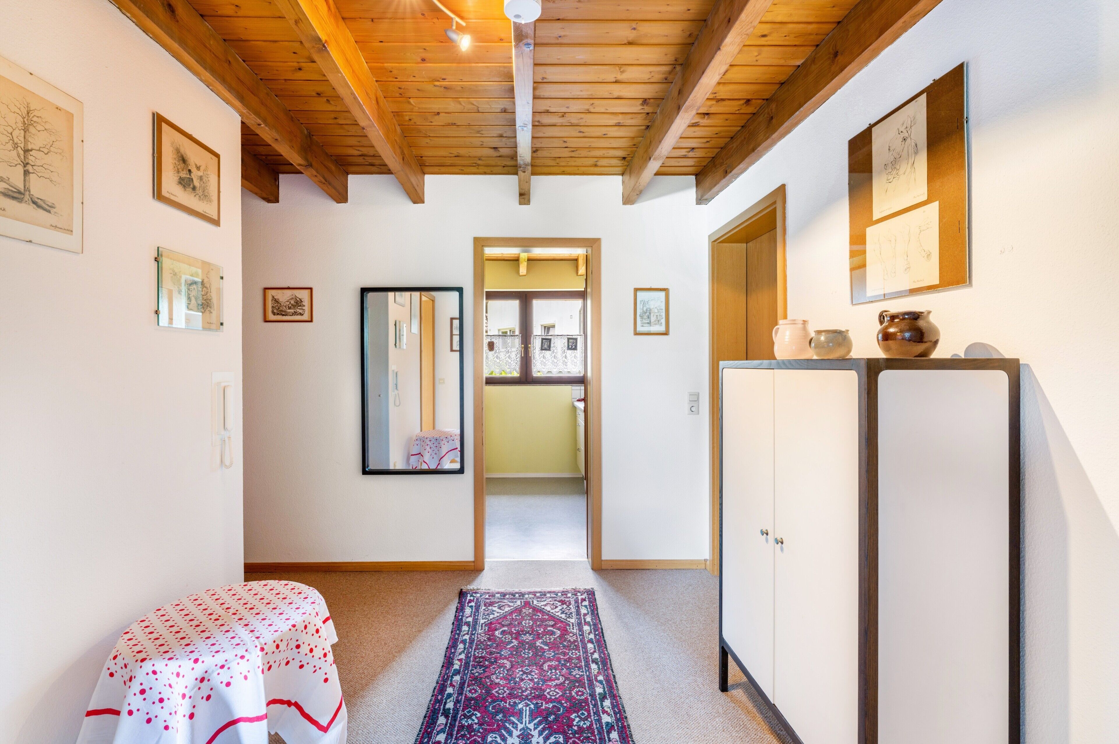 Ein Flur mit Teppichmuster, Holzbalkendecke, einem kleinen Tisch mit Punkte-Tischdecke und einem Schrank mit Dekorationen.