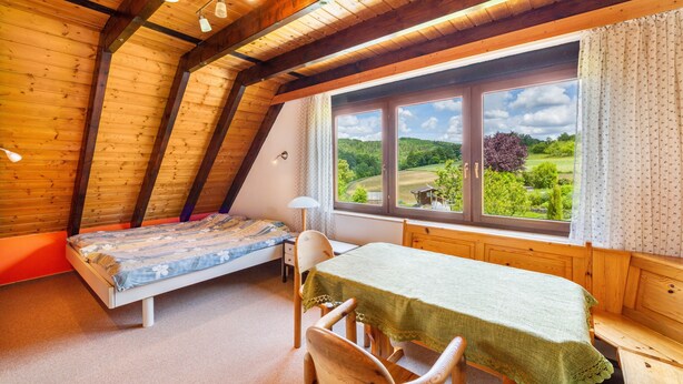 Ein Zimmer mit schrägen Wänden, einem Bett, einem Tisch mit Tischdecke, einem Stuhl und einem Fenster mit Blick auf Grünflächen.