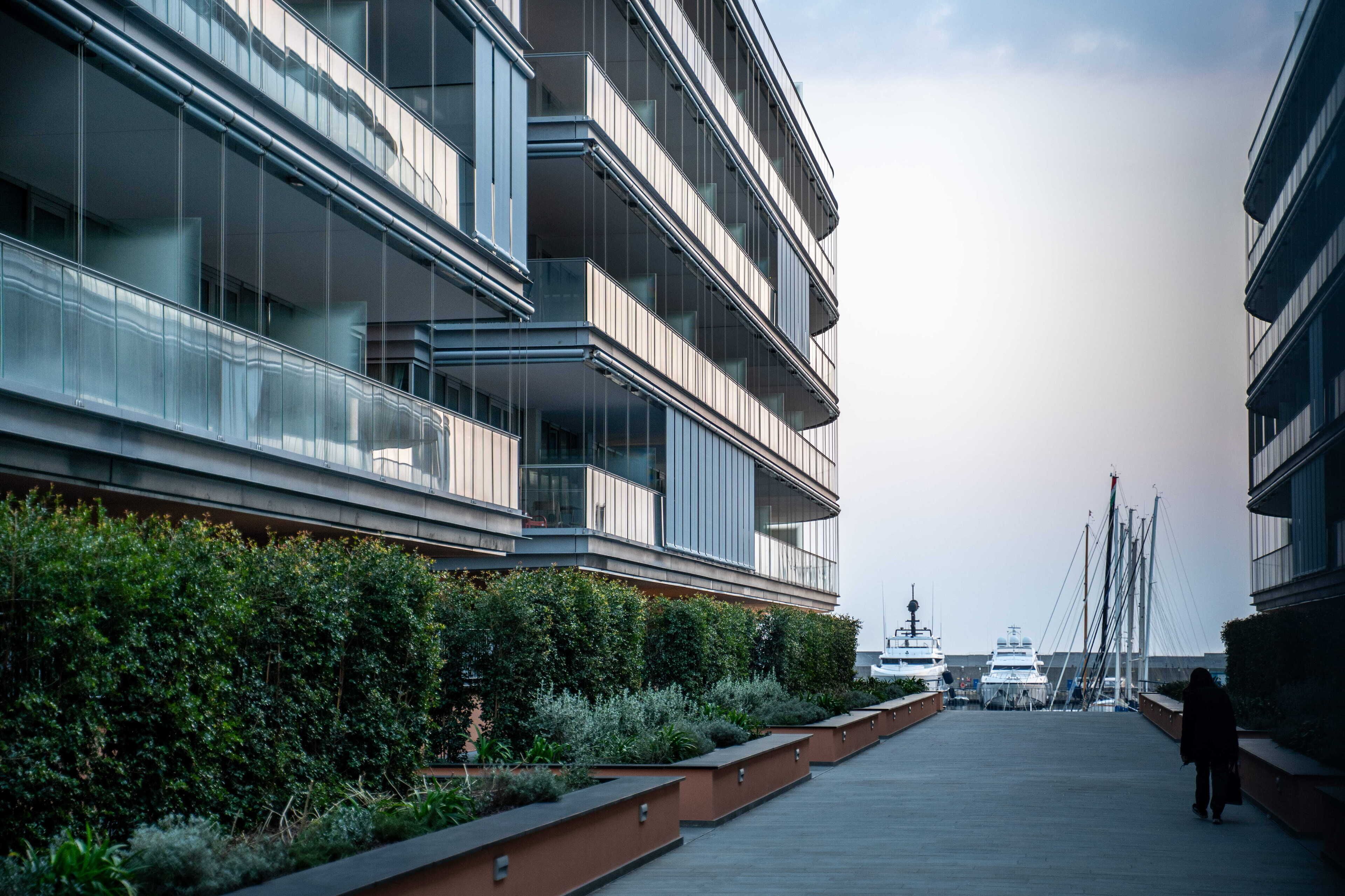 A modern multi-story building with balconies, a waterfront view, and a marina in the background.