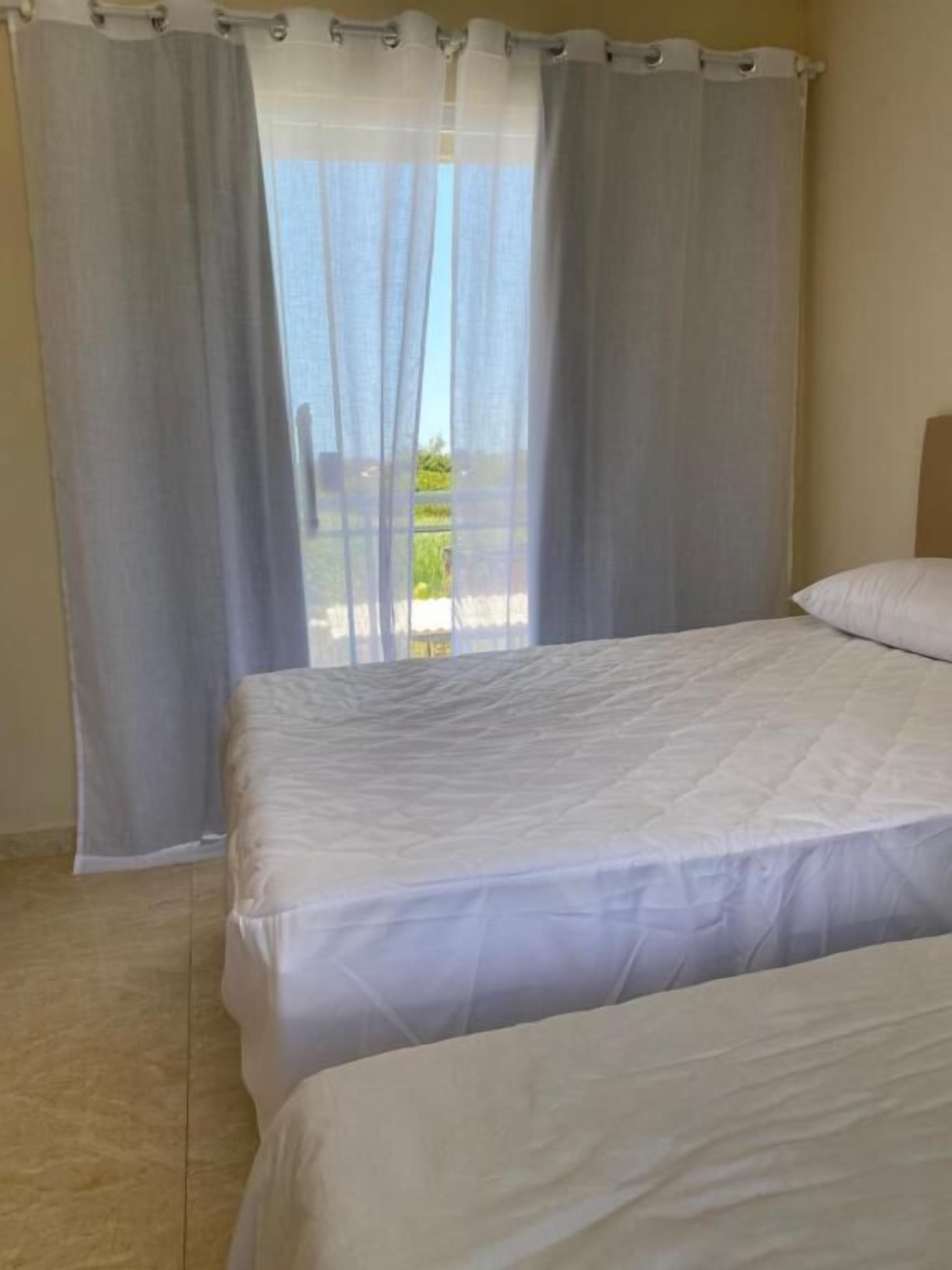 A bedroom with two beds, a view of the outdoors through sheer curtains, and a tiled floor.