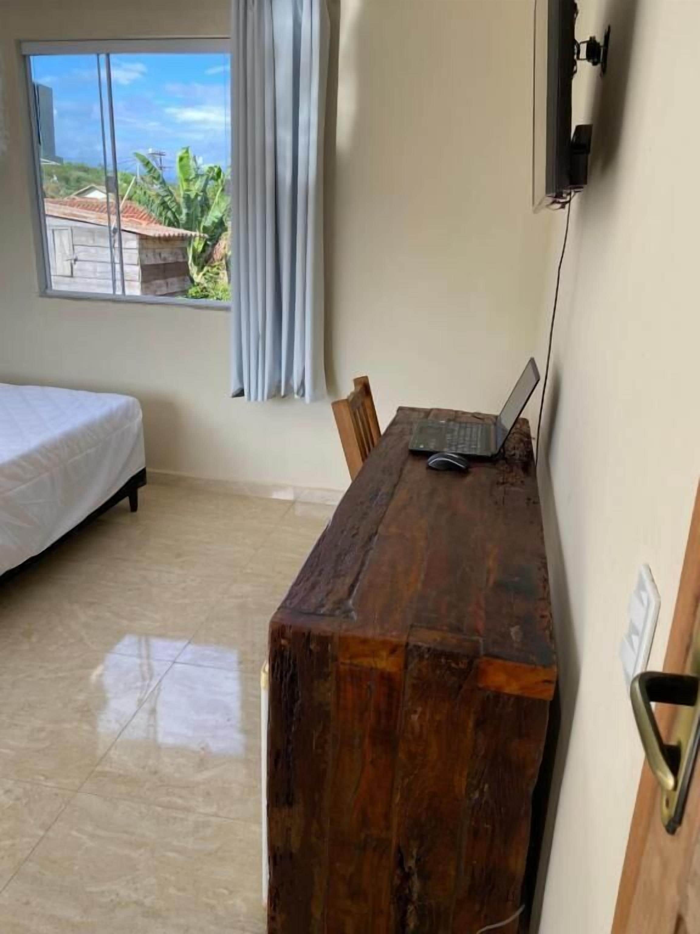 Una habitación con cama, un escritorio de madera con una computadora portátil, una ventana con cortinas y vistas a edificios y áreas verdes.
