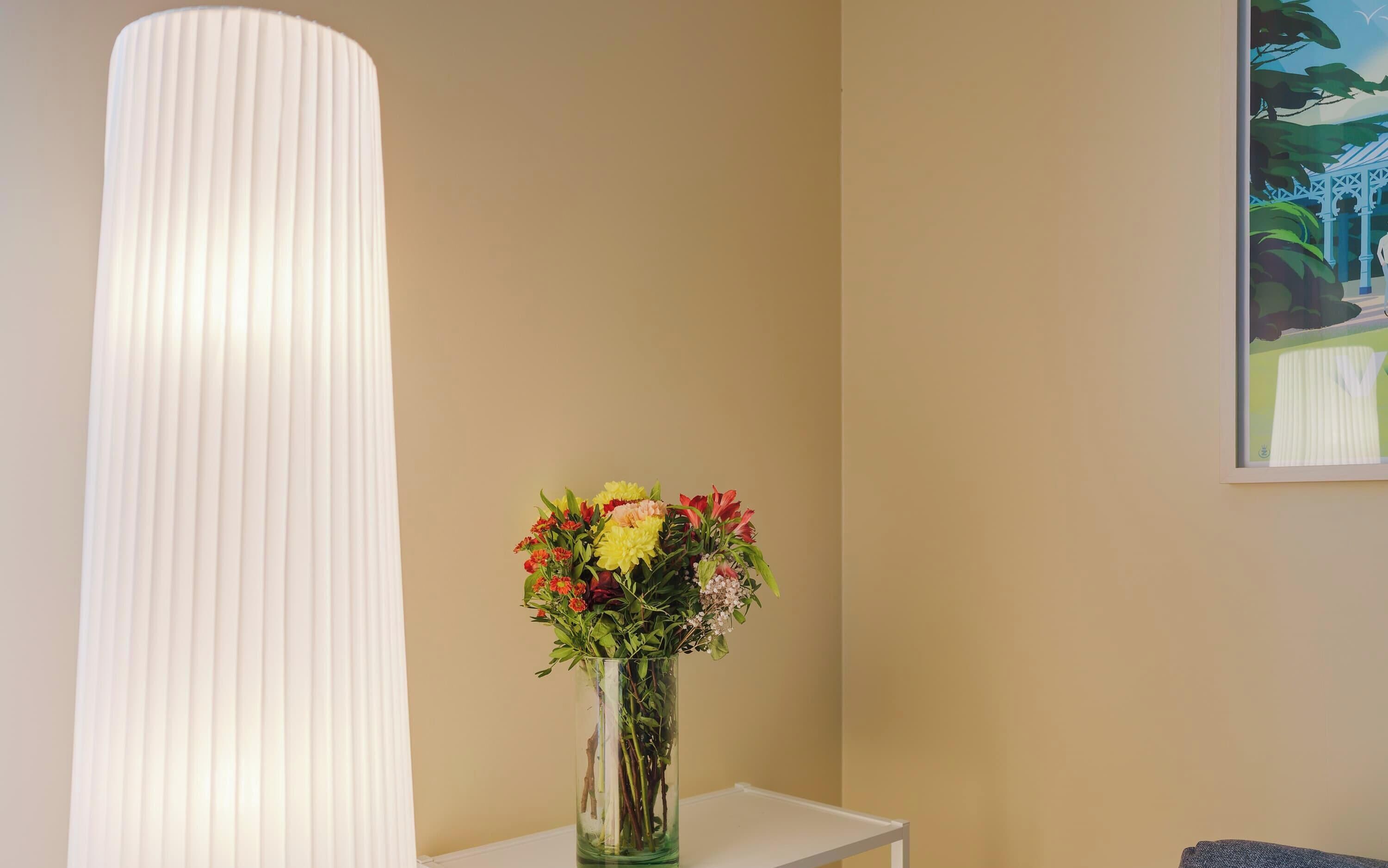 Une pièce avec une grande lampe blanche à motifs en relief, un vase en verre transparent rempli de fleurs et un tableau encadré accroché au mur.