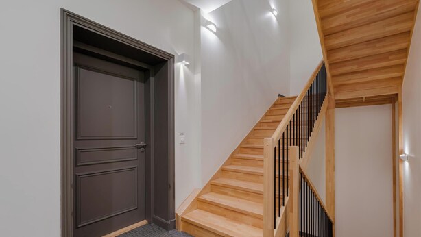 Eine Treppe mit hölzernen Stufen und schwarzen Spindeln, eine dunkle Holztür und eingelassene Wandleuchten.