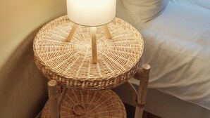 Ein Bett mit weißer Bettwäsche, ein runder Weidenholz-Nachttisch mit Lampe und ein Korbwäscher.