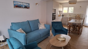 A living room with a blue sofa, a wooden coffee table, and a dining area with a table and chairs. - Residence close to beaches with terrace and garden (Saint-Coulomb)