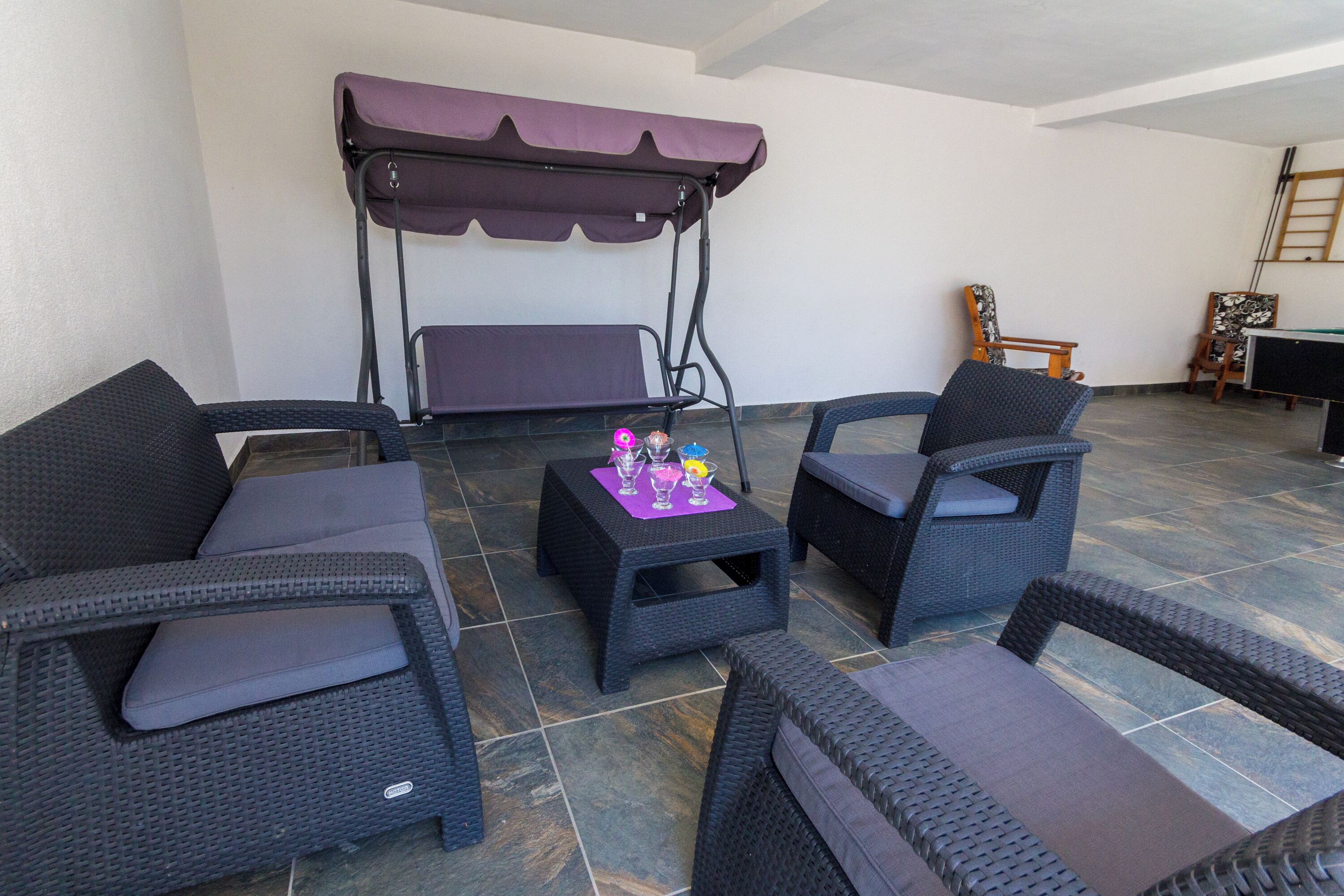 A covered outdoor seating area with wicker furniture, a swing, and a small table with drinks.