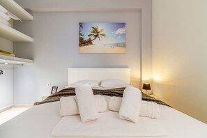 Ein ordentlich bezogenes Bett mit weißen Leinen und Handtüchern, ein Kopfteil, ein gerahmtes Bild von einer Strandansicht und eine Nachttischlampe.