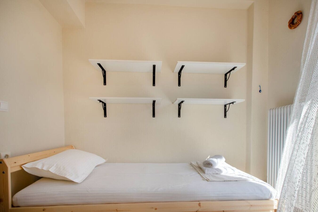 Una cama individual con cabecera de madera, almohada blanca y una toalla enrollada sobre la cama. Hay cuatro estantes fijos en la pared arriba.