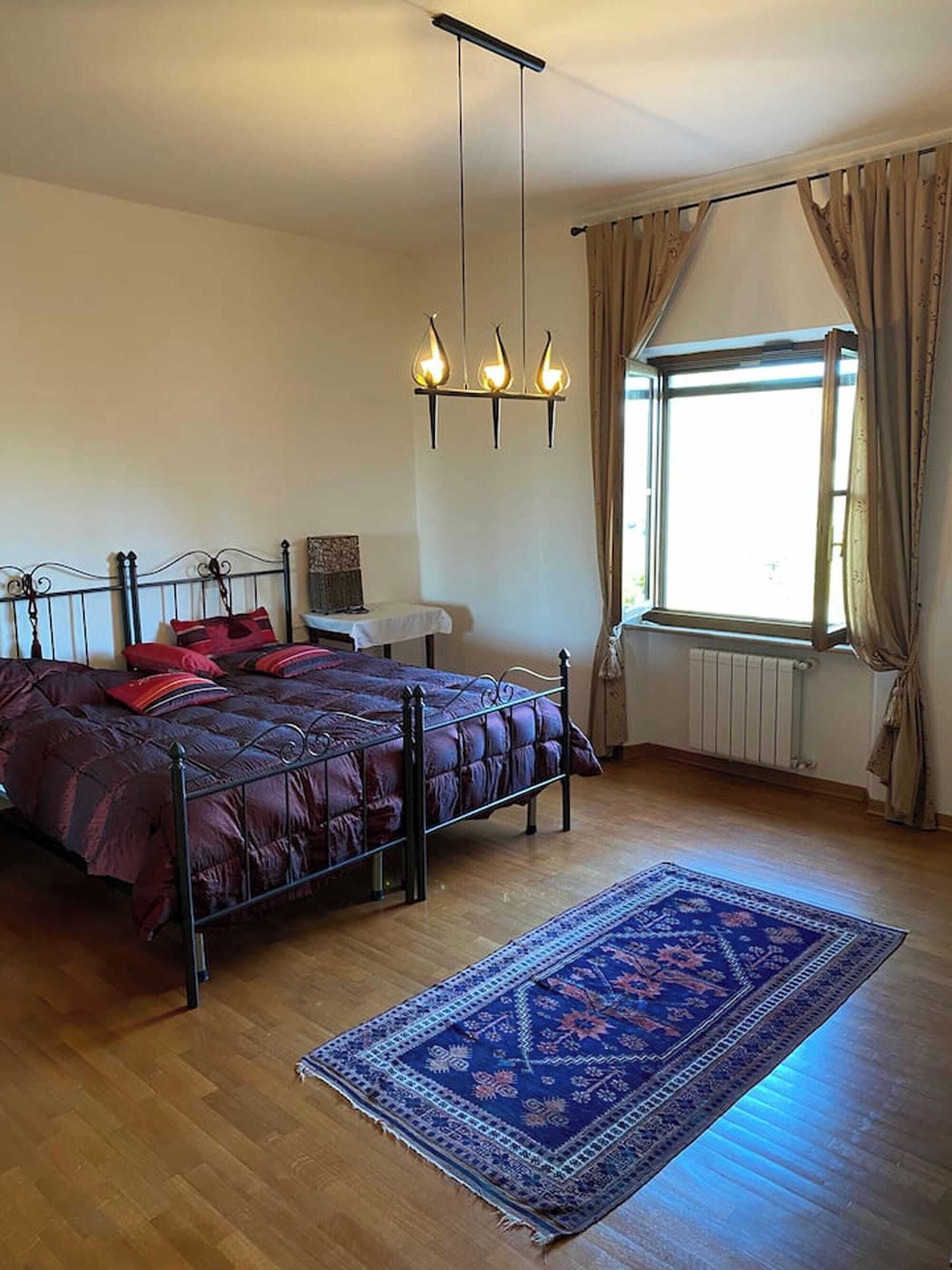Un dormitorio con una cama, una lámpara colgante, una ventana con cortinas y una alfombra estampada.