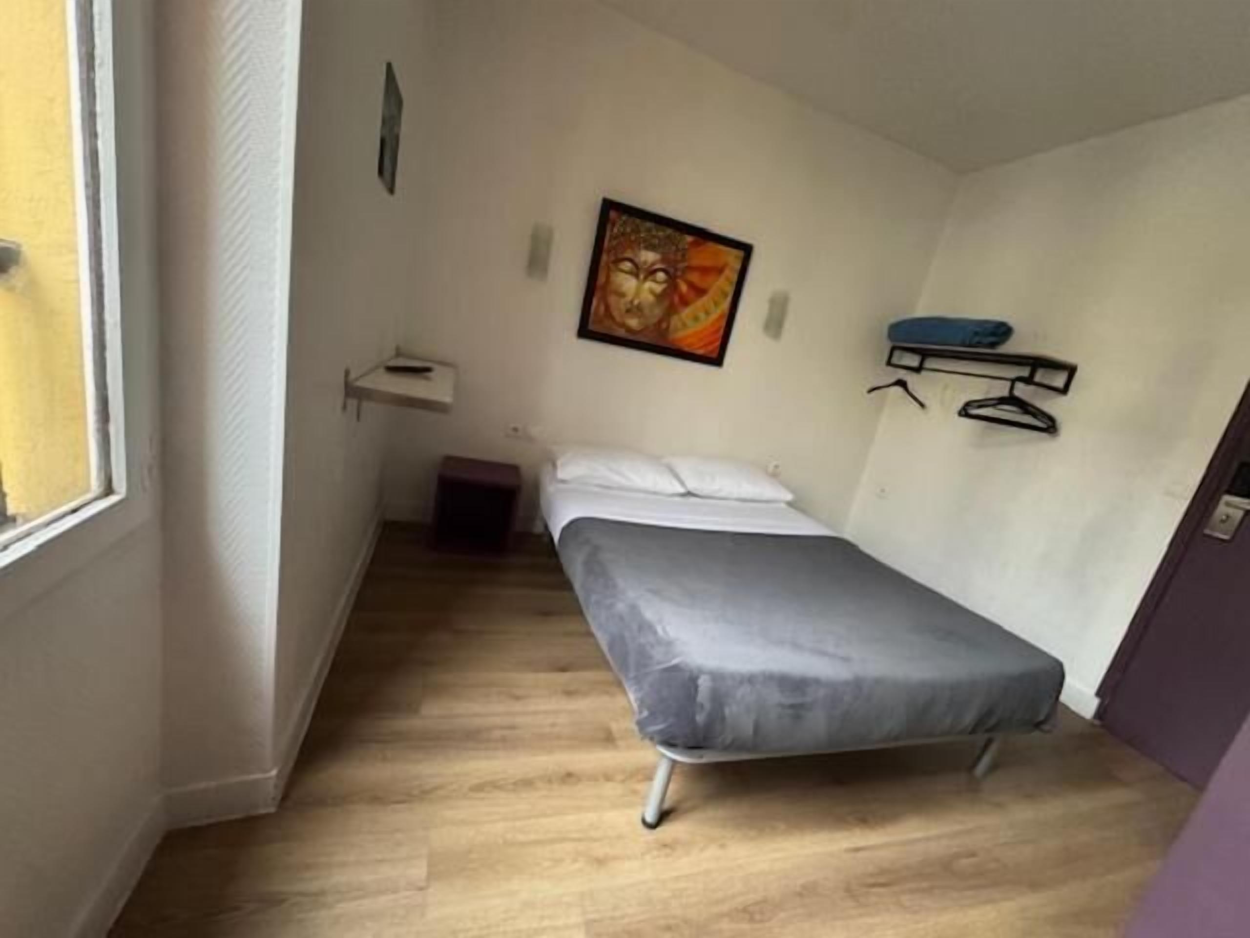 A small, single-bed room with a wooden floor, a bedside table, a wall-mounted shelf, and a window.