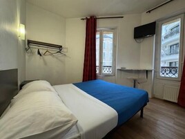 Quarto de hotel com uma cama, uma televisão presa à parede e uma janela com vista para prédios.