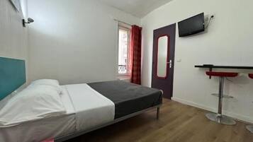 A single bed with a headboard, a small table with a chair, a television mounted on the wall, and a door with a mirror.
