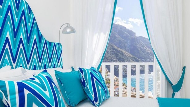 Een slaapkamer met een bed, kussens, een lamp en uitzicht op zee en bergen.