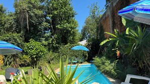 Ein Pool mit Liegestühlen und Sonnenschirmen, umgeben von üppiger Vegetation und einer Ziegelmauer.