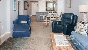A living room with a blue sofa, a black leather chair, and a coffee table. There is a dining area with a table and chairs in the background. - Big Surf 1E 2D - Gulf View Beach Pool and Fun (Gulf Shores)