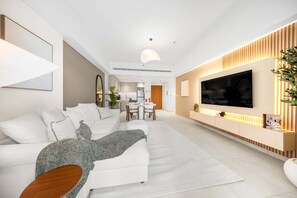 A modern living room with a sofa, a TV, and a dining area in the background. - Free Housekeeping Visits During Your Stay - Stayshort - Modern 1BR Retreat in MJL Sleeps 4 (Dubai)