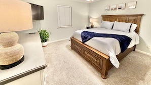 A bedroom with a bed, a ceiling fan, a TV, a lamp, a plant, and a window.