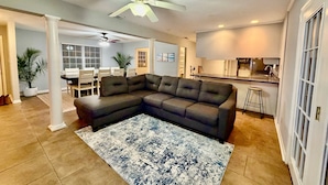 A living room with a sectional sofa, a dining area with a table and chairs, a kitchen with a bar, and a ceiling fan.