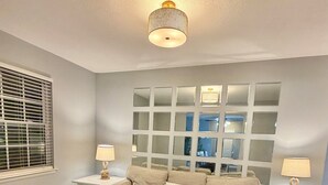 A living room with a sofa, two side tables with lamps, a ceiling light fixture, and a large window with blinds.